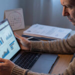 Un homme concentré examine des graphiques financiers sur son ordinateur portable, un calendrier et des documents sont sur le bureau. Il semble préoccupé par un retard de paiement.