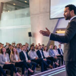 A charismatic speaker in a dark suit gestures on a futuristic, blue-lit stage to an engaged, diverse audience in a modern hall.