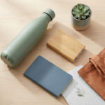 Flat lay of modern sustainable items: green water bottle, bamboo power bank, blue & seed paper notebooks, succulent, and brown tote bag on wood.