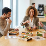 Diverse colleagues enjoy fresh, packaged corporate meals in a bright, modern office break room. One opens a salad, another looks at salmon.